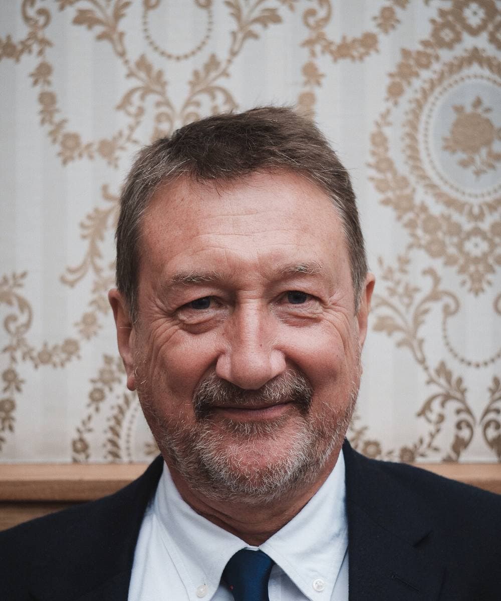 Steven Knight, grey-haired and bearded, in a white shirt, blue tie and dark jacket, smiling in front of patterned wallpaper