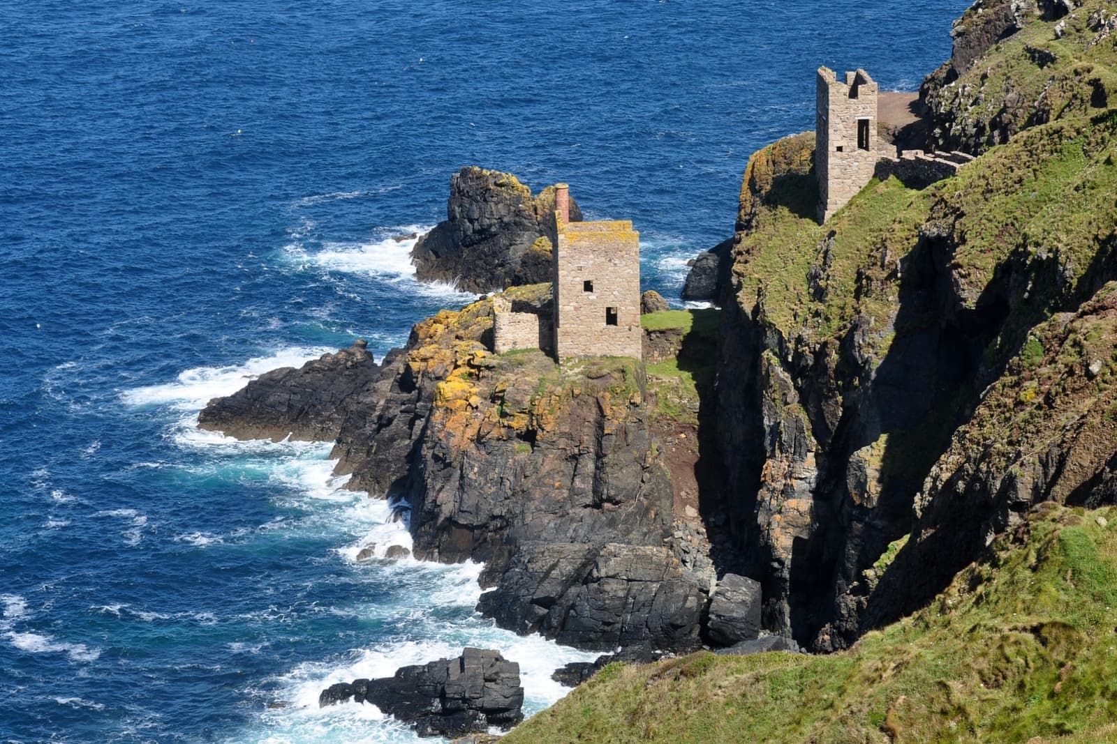 Two ruined engine houses on a rocky ledge at the foot of green cliffs above a dark blue sea