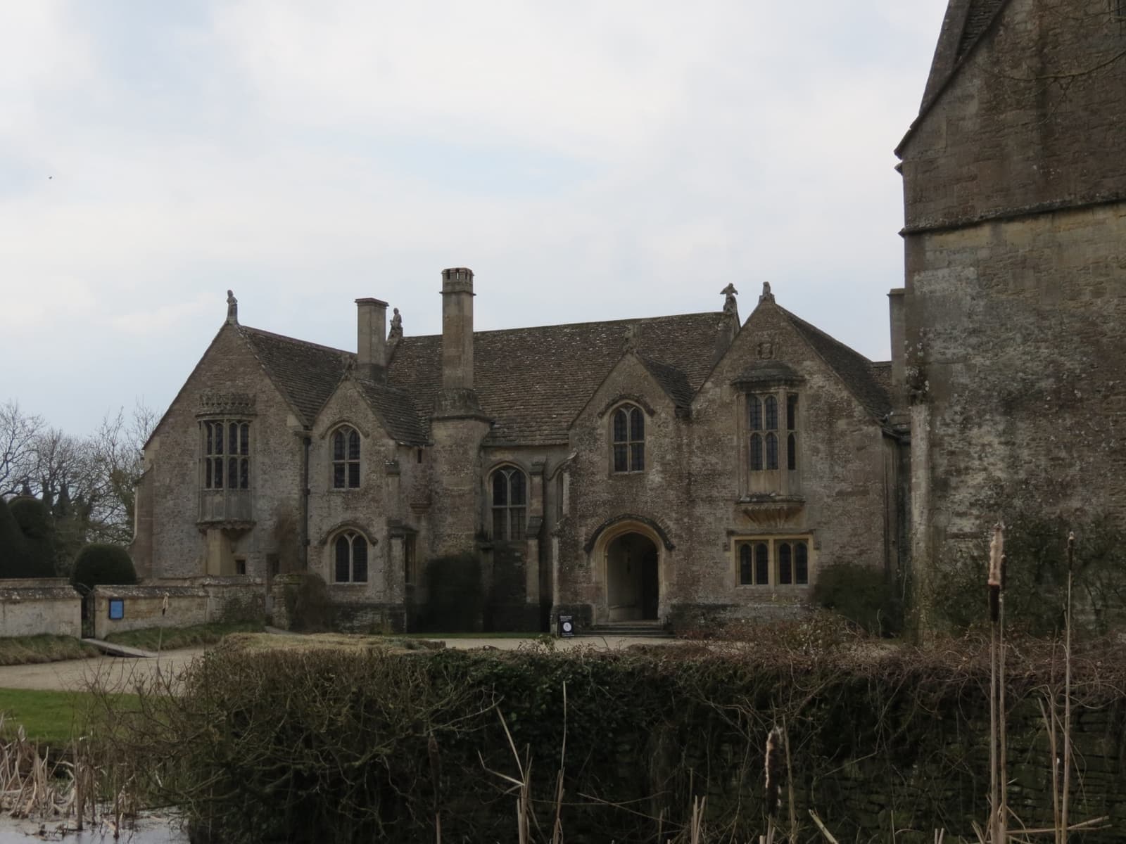 A gabled stone manor house with tall windows behind a moat hedge