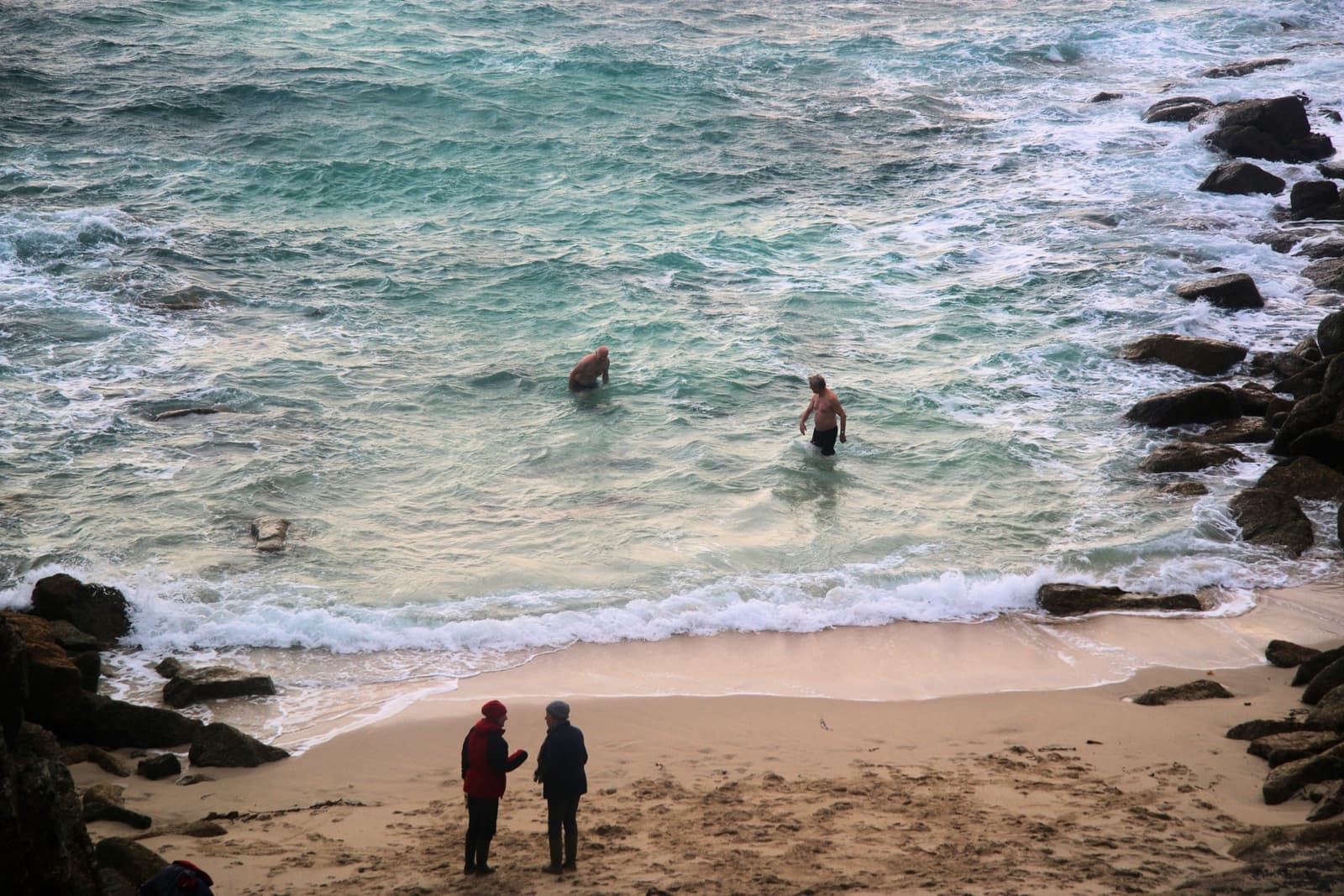 Two people swimming in green surf in a small cove while two others watch from the sand