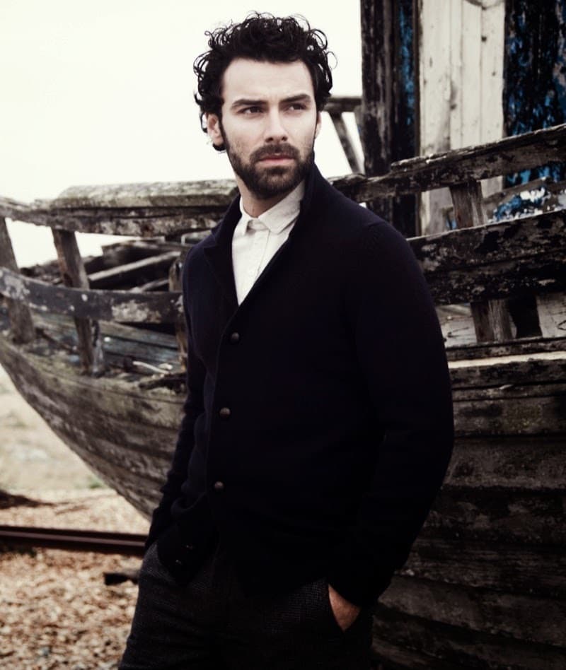 Aidan Turner, bearded, in a dark coat, standing in front of an old wooden boat