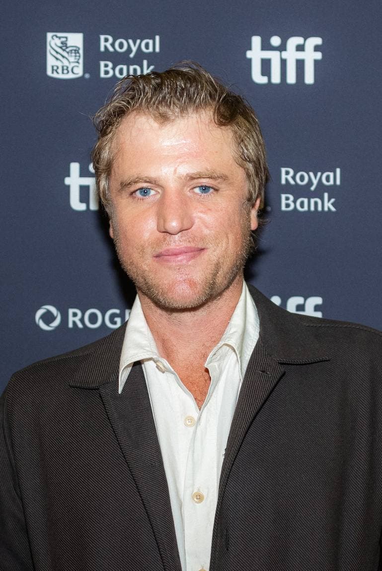 Johnny Flynn in a dark jacket and open white shirt, looking at the camera