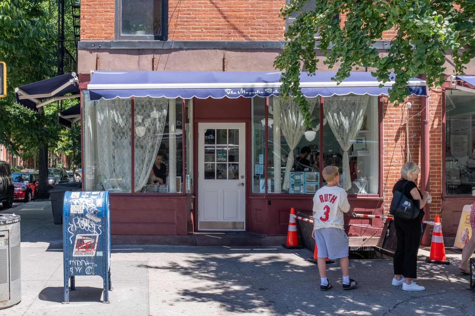 A small corner bakery with a blue scalloped awning and lace curtains, people at the door