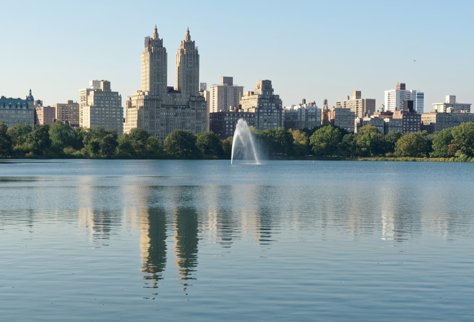 A wide still reservoir with a fountain plume and twin-towered apartment buildings beyond the trees
