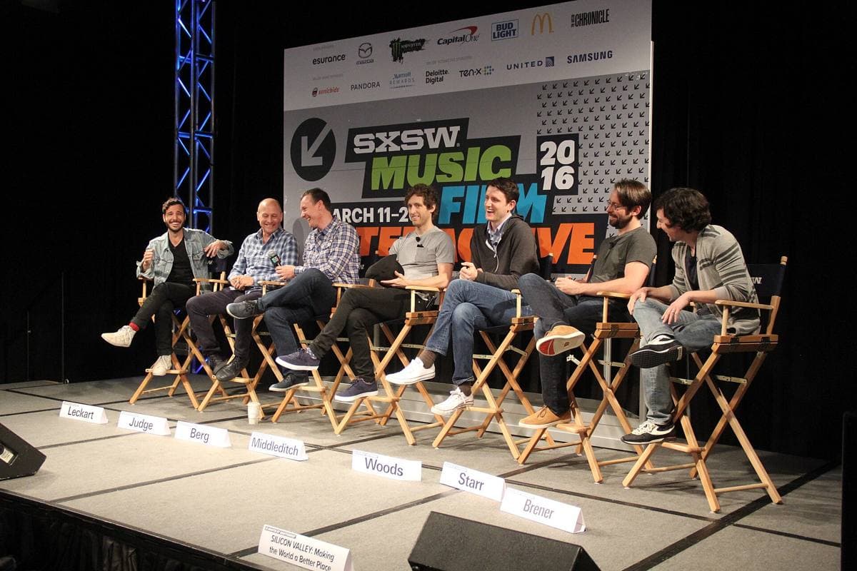 Seven men seated in a row on a panel stage with microphones