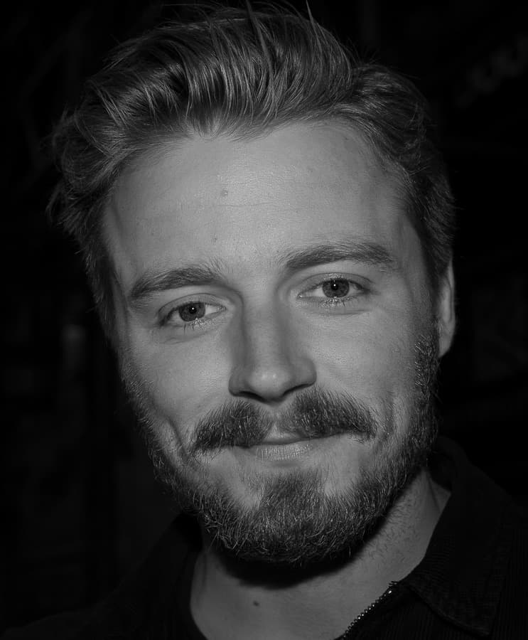 Jack Lowden in black and white, bearded, smiling slightly