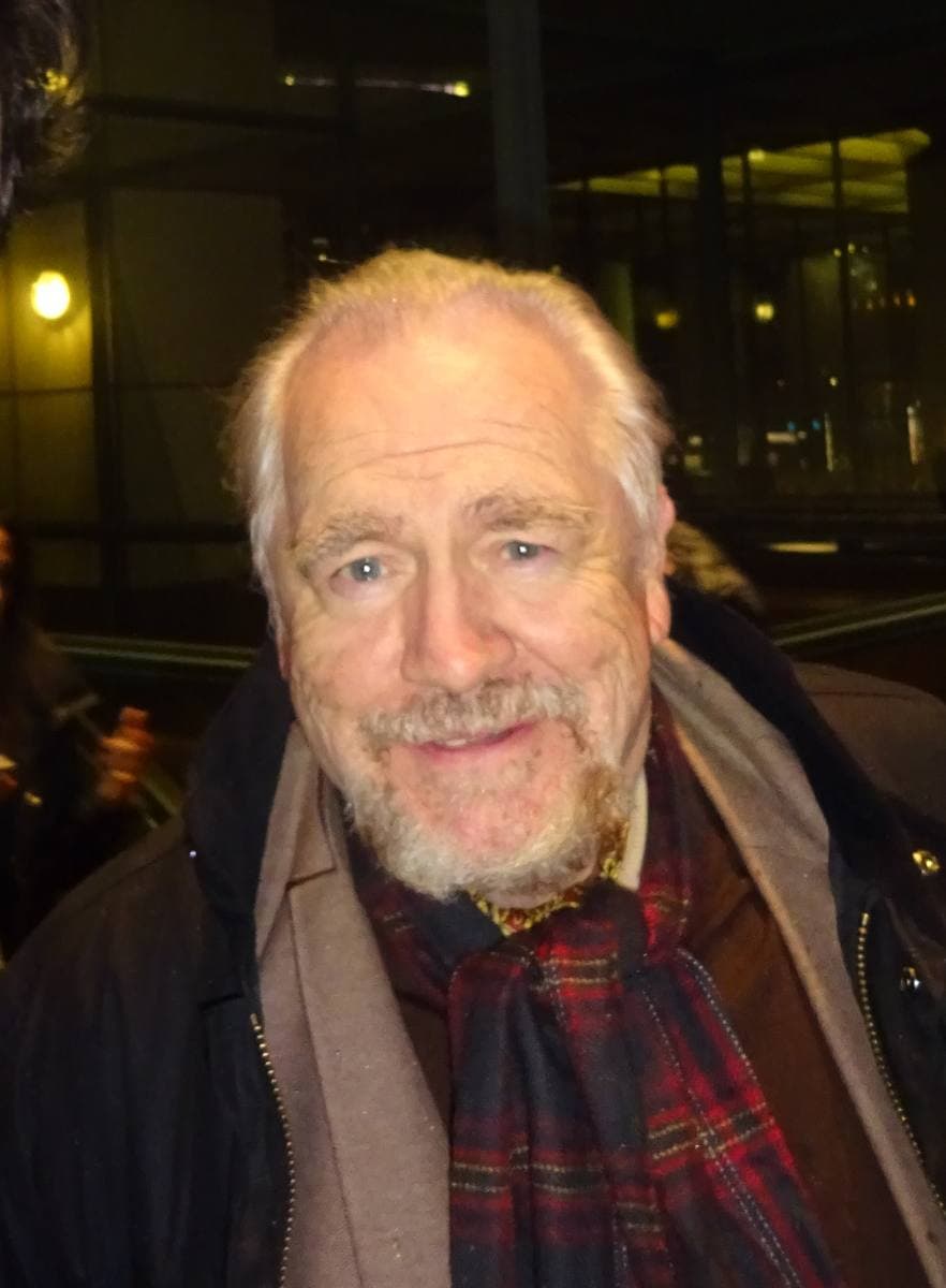 Brian Cox with a white beard, in a dark coat and tartan scarf, at night
