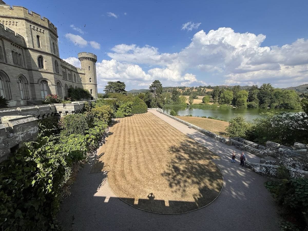 A gravel terrace beside Eastnor Castle’s round tower, looking down to the lake under summer cloud