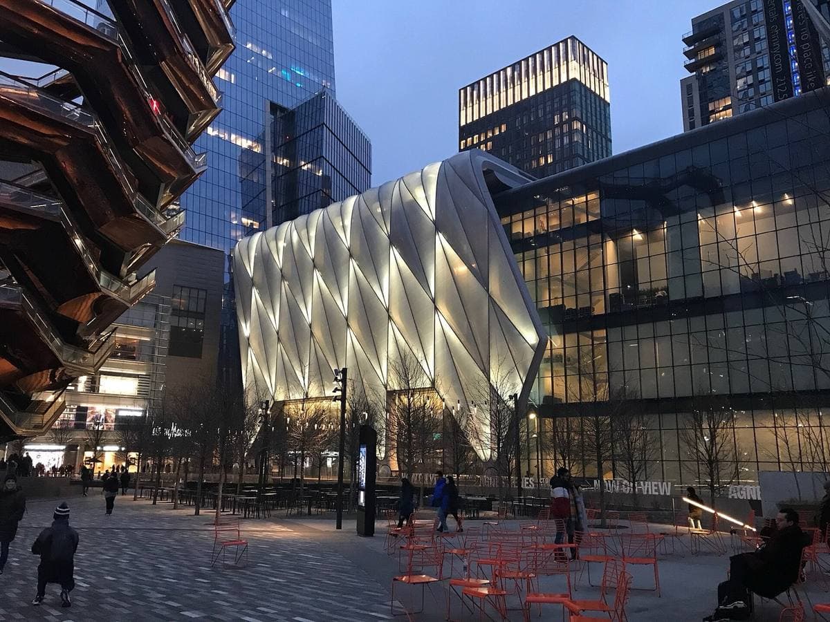 The quilted, lit shell of The Shed at dusk, with red café chairs on the plaza