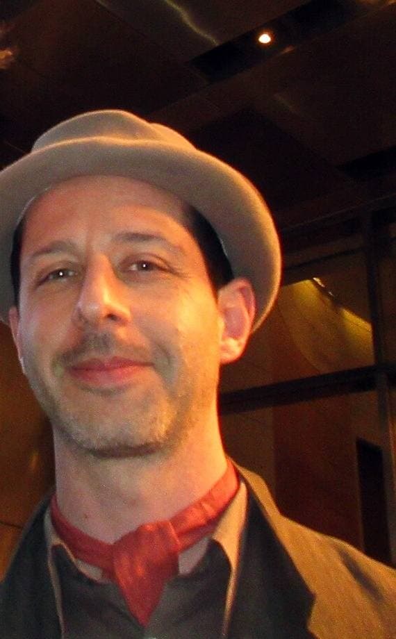 Jeremy Strong in a grey trilby, red neckerchief and tweed jacket, smiling slightly