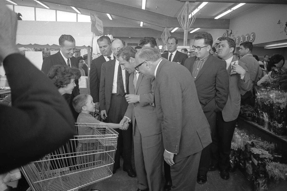 Men in suits crowd round a visitor between the shelves and trolleys of a supermarket, 1962
