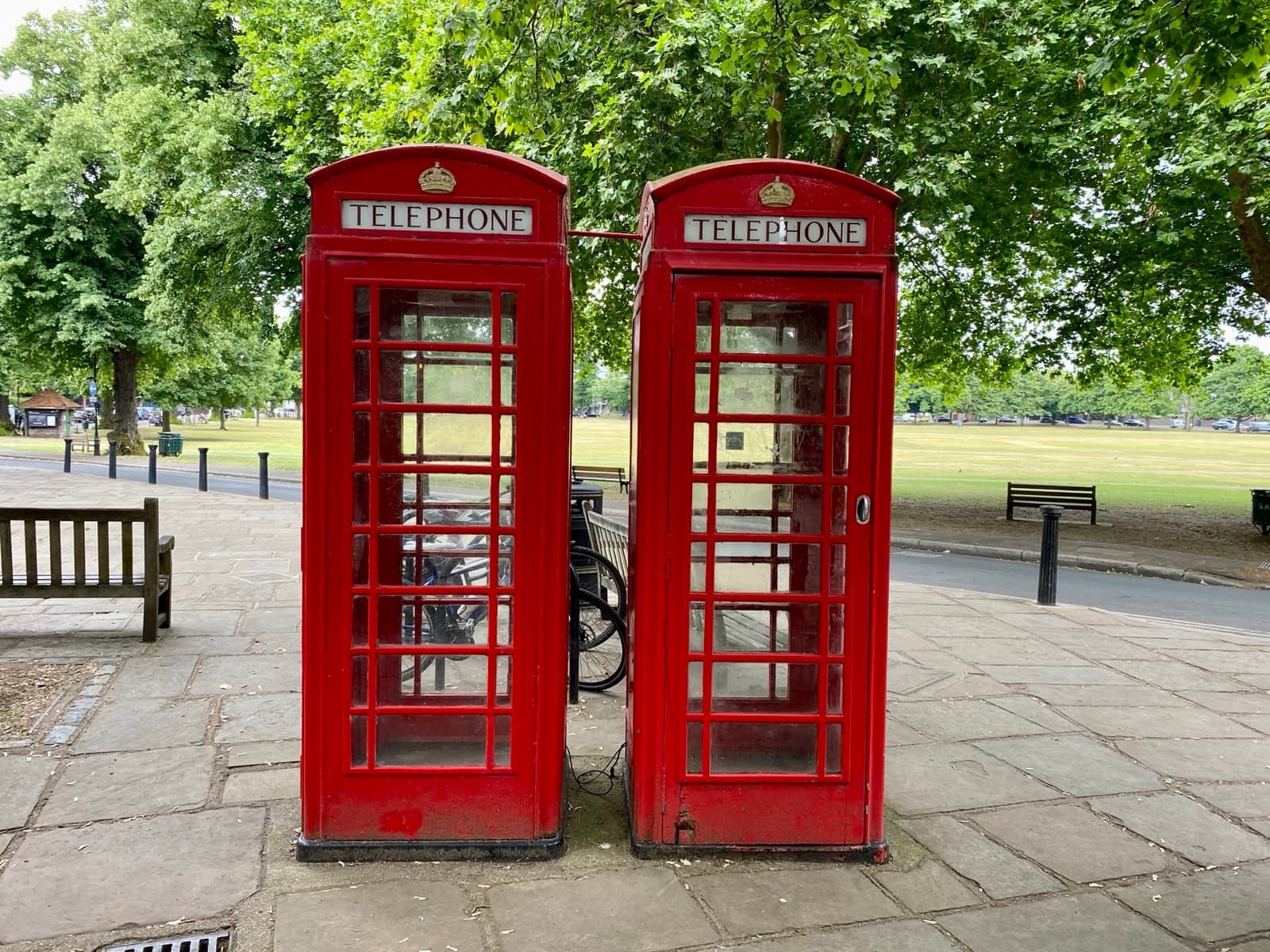 Two red K6 telephone boxes side by side on paving at the edge of a tree-lined green