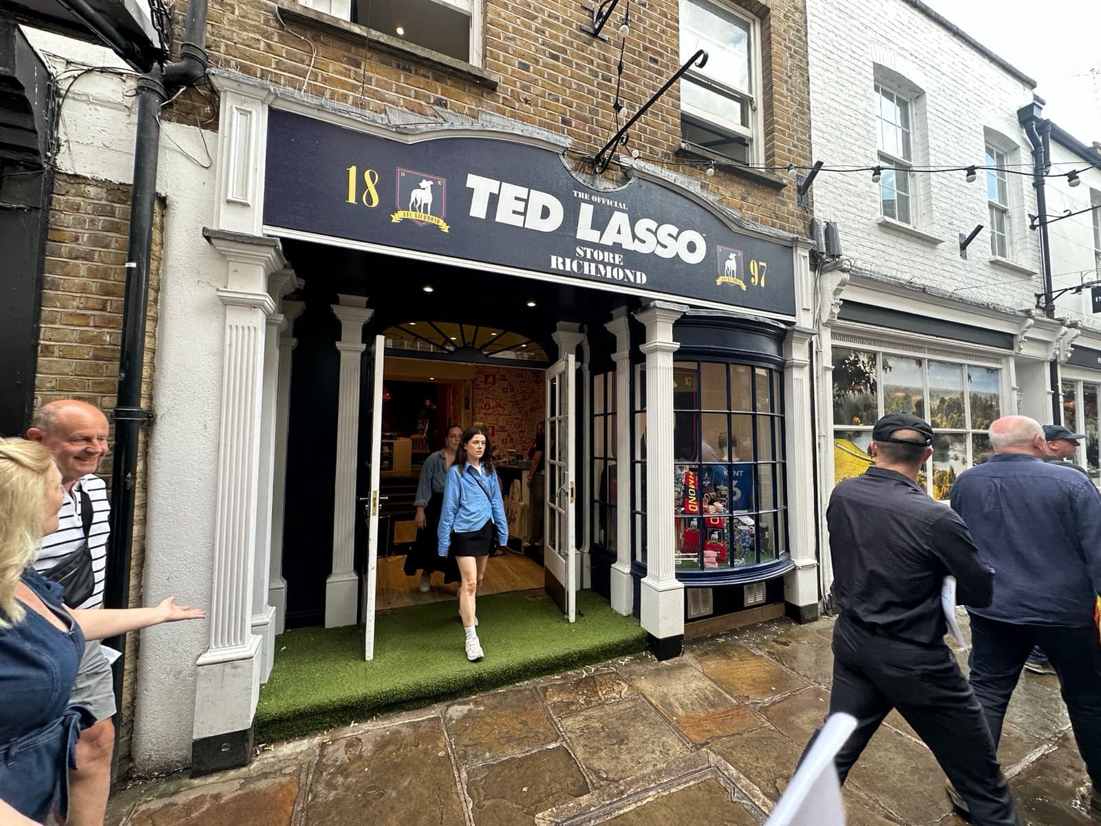 A navy shopfront with a sign reading Ted Lasso and people walking past on the paving