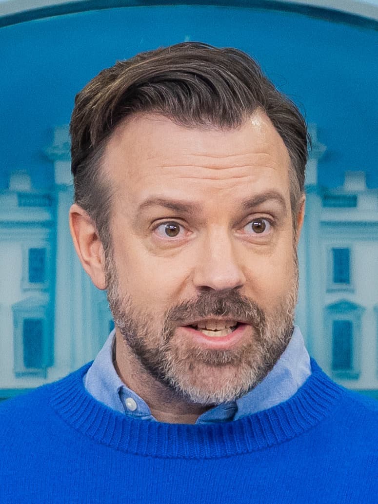 Jason Sudeikis in a bright blue jumper over a pale blue shirt, mid-sentence