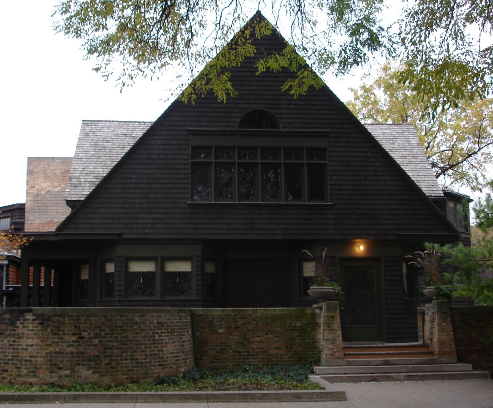 A shingled house with a steep triangular gable and a low brick wall in front