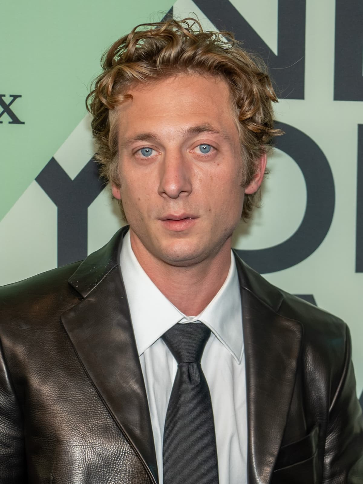 Jeremy Allen White, curly fair hair, in a black leather jacket and dark tie, looking at the camera