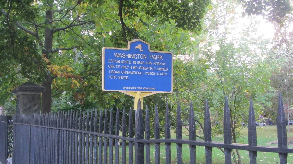 A fenced square of lawn and trees ringed by brownstone row houses