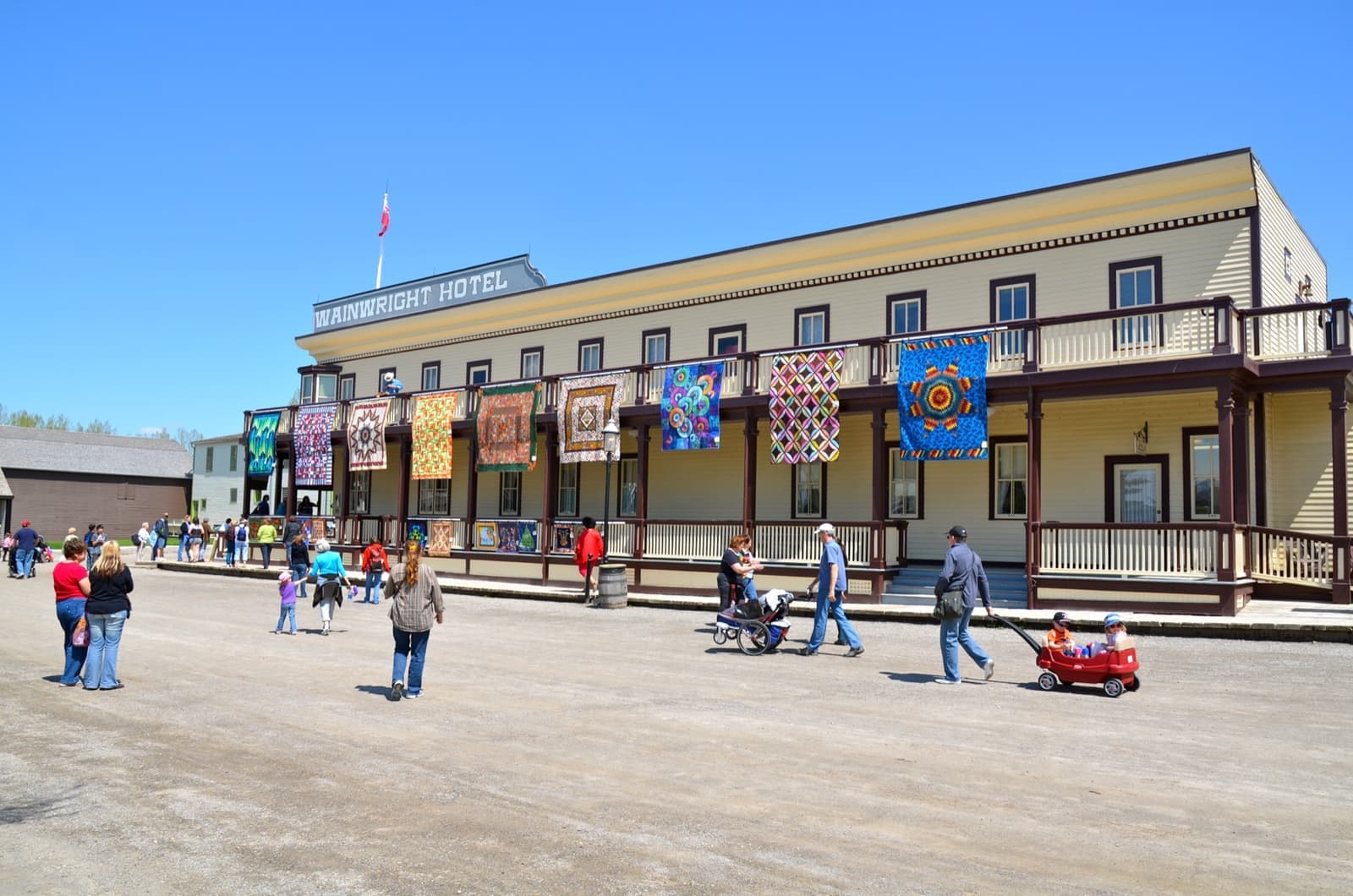 A long two-storey frontier hotel with a balcony and flags on a dirt street, people strolling