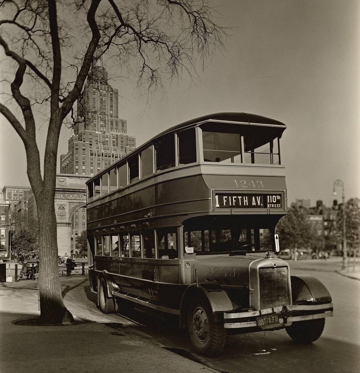 A black-and-white photograph of a double-decker bus under bare trees, a marble arch and a tall apartment tower behind it