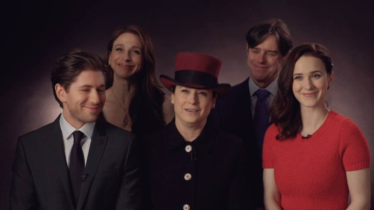 Five people posed close together against a dark backdrop: a woman in a red jumper on the right, a woman in a red-and-black top hat in the centre