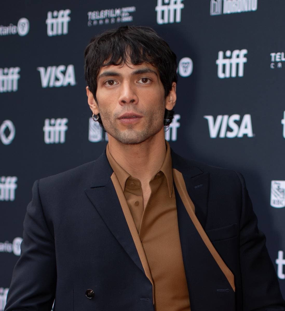 Diego Calva with dark curly hair, in a dark suit