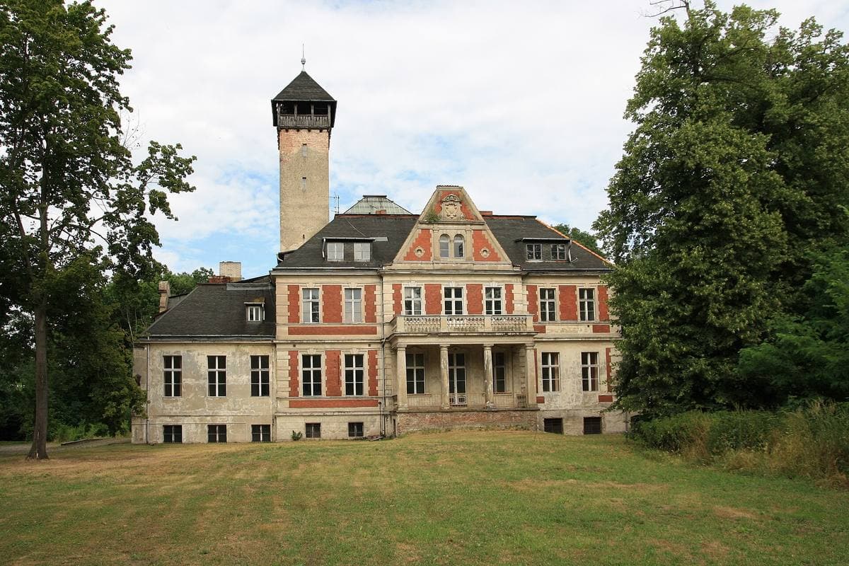 A grand pale manor house with a tower behind a fence and trees