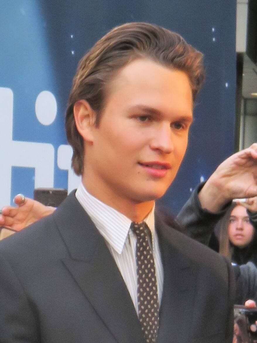 Ansel Elgort in a dark suit and tie, looking to one side
