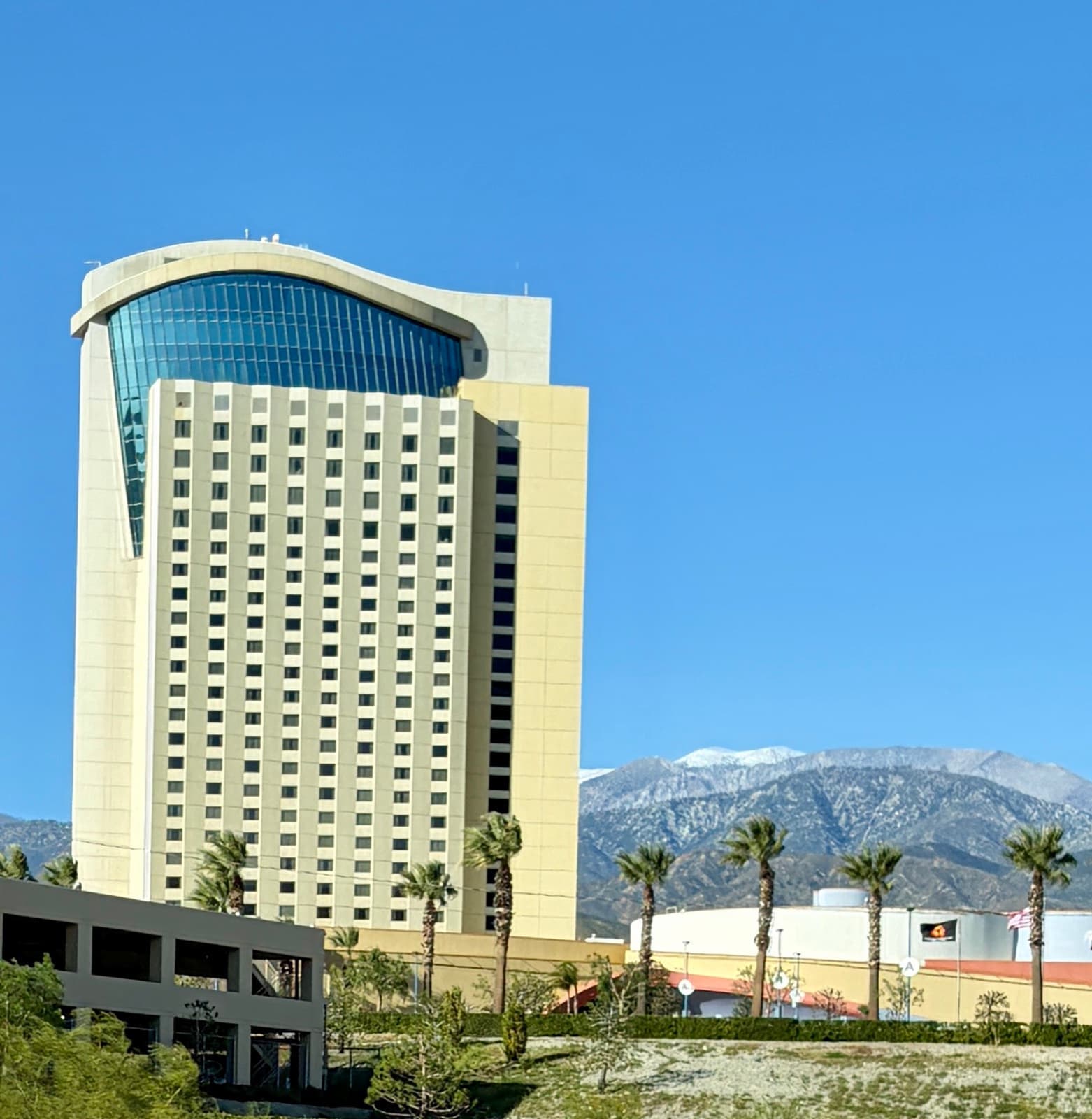 Morongo hotel tower and grounds beneath blue sky with snowy mountains beyond.