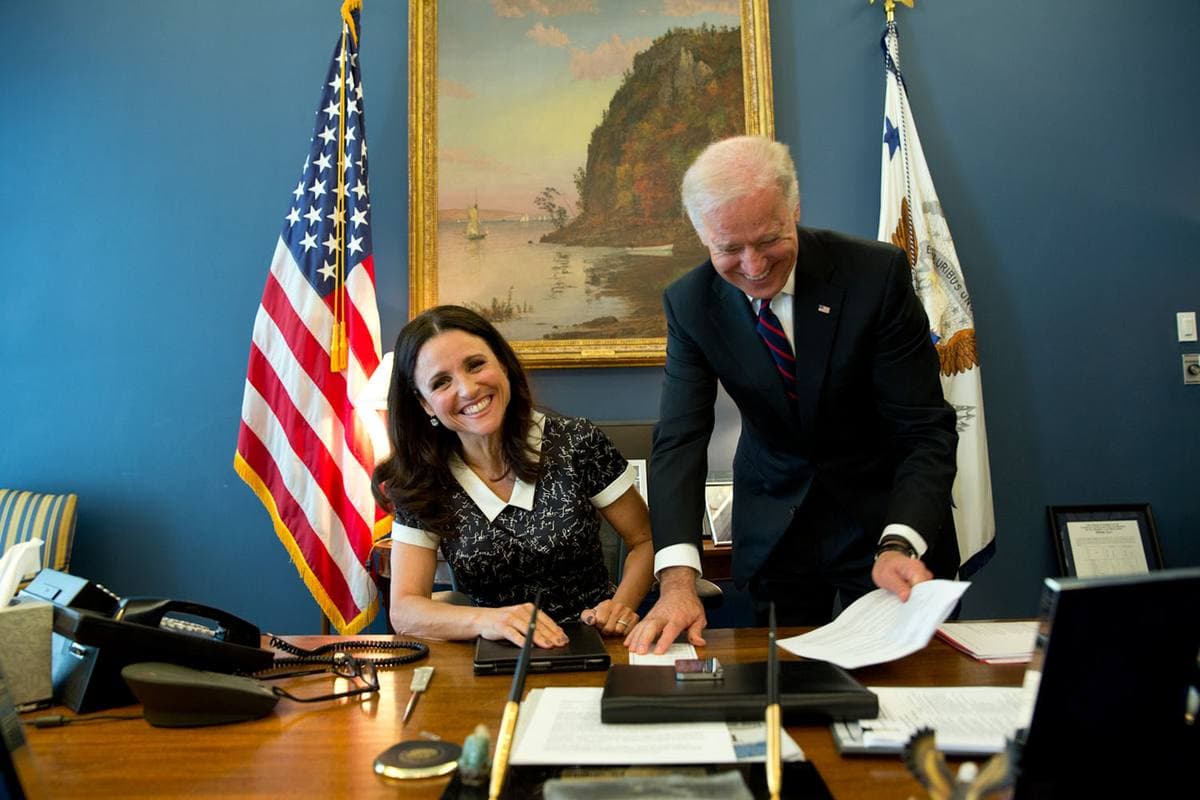 Julia Louis-Dreyfus seated at a desk between flags, smiling, as Joe Biden leans over to show her a sheet of paper