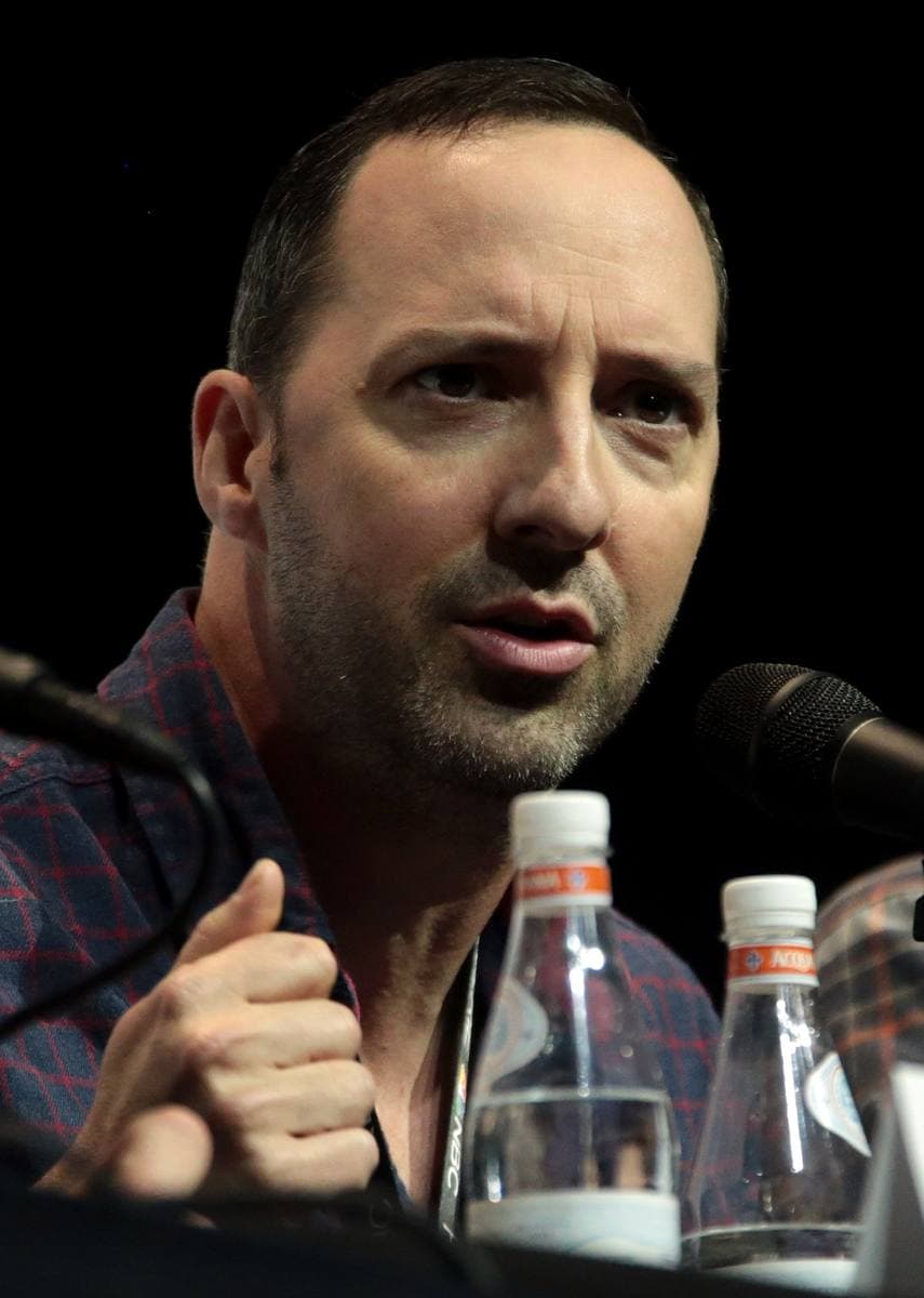 Tony Hale with a short beard speaking at a microphone, water bottles in front of him
