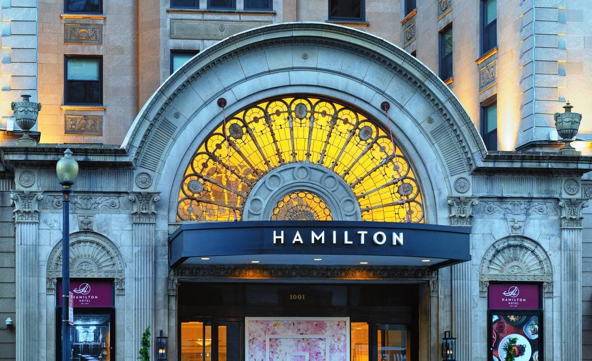 A stone arched hotel entrance with a gold stained-glass fan window and a blue canopy reading Hamilton