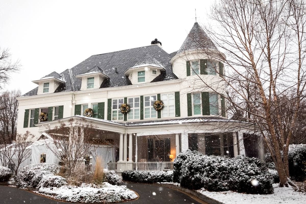 A white Queen Anne house with a turret and a wide porch in light snow, wreaths in the windows