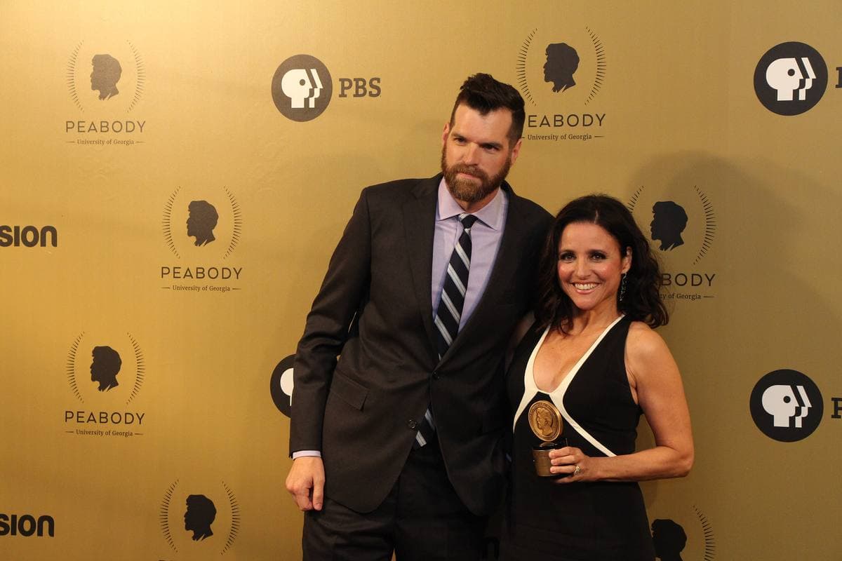 Timothy Simons in a dark suit and Julia Louis-Dreyfus in a black dress holding an award in front of a gold backdrop