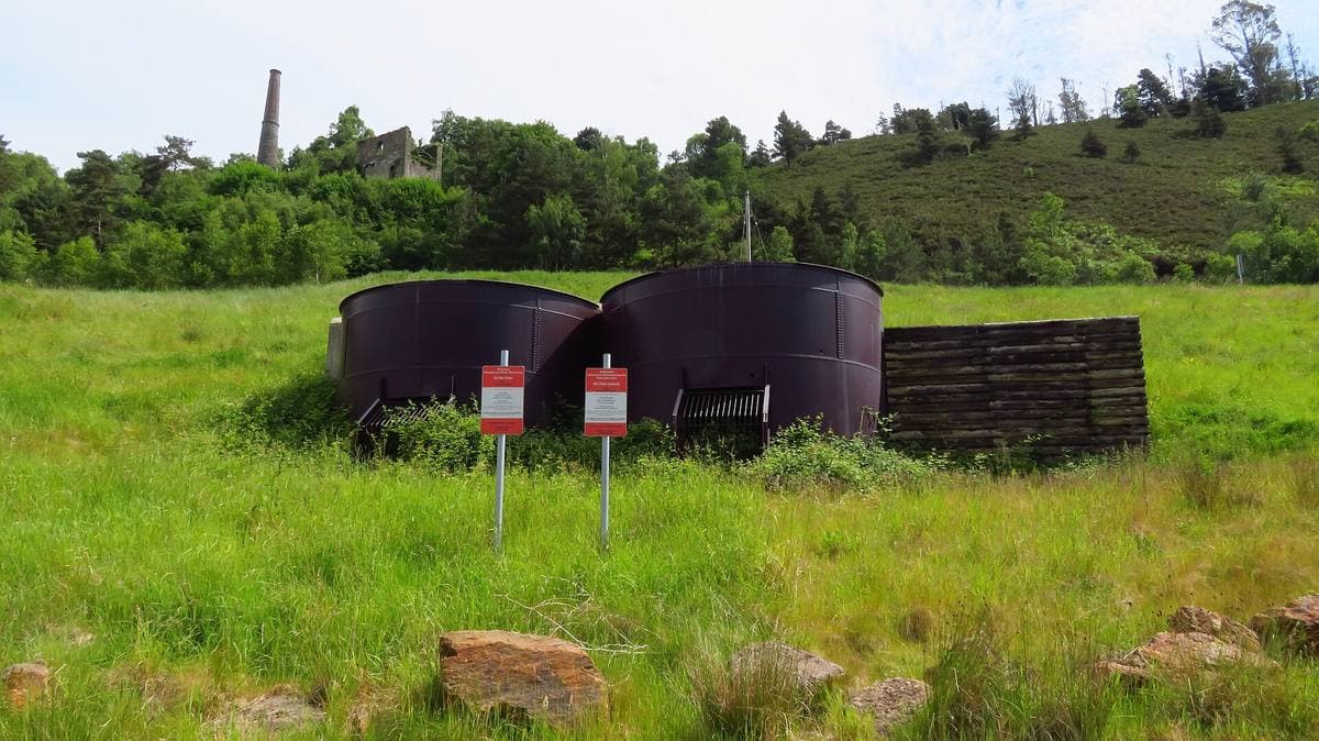 Two rusty round ore bins in a meadow, a stone chimney on the wooded hill behind