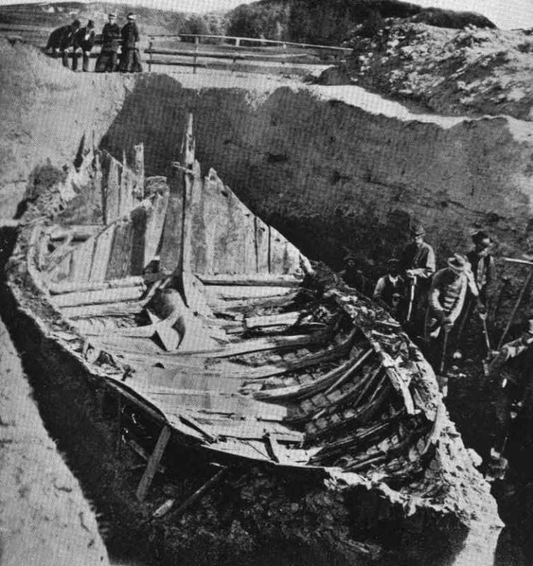 An old photograph of a ship’s hull lying in a dug-out mound, people watching from above