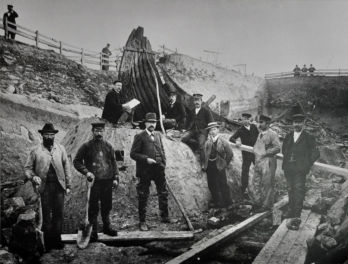 Men in hats standing in a trench in front of the curved prow of a buried ship