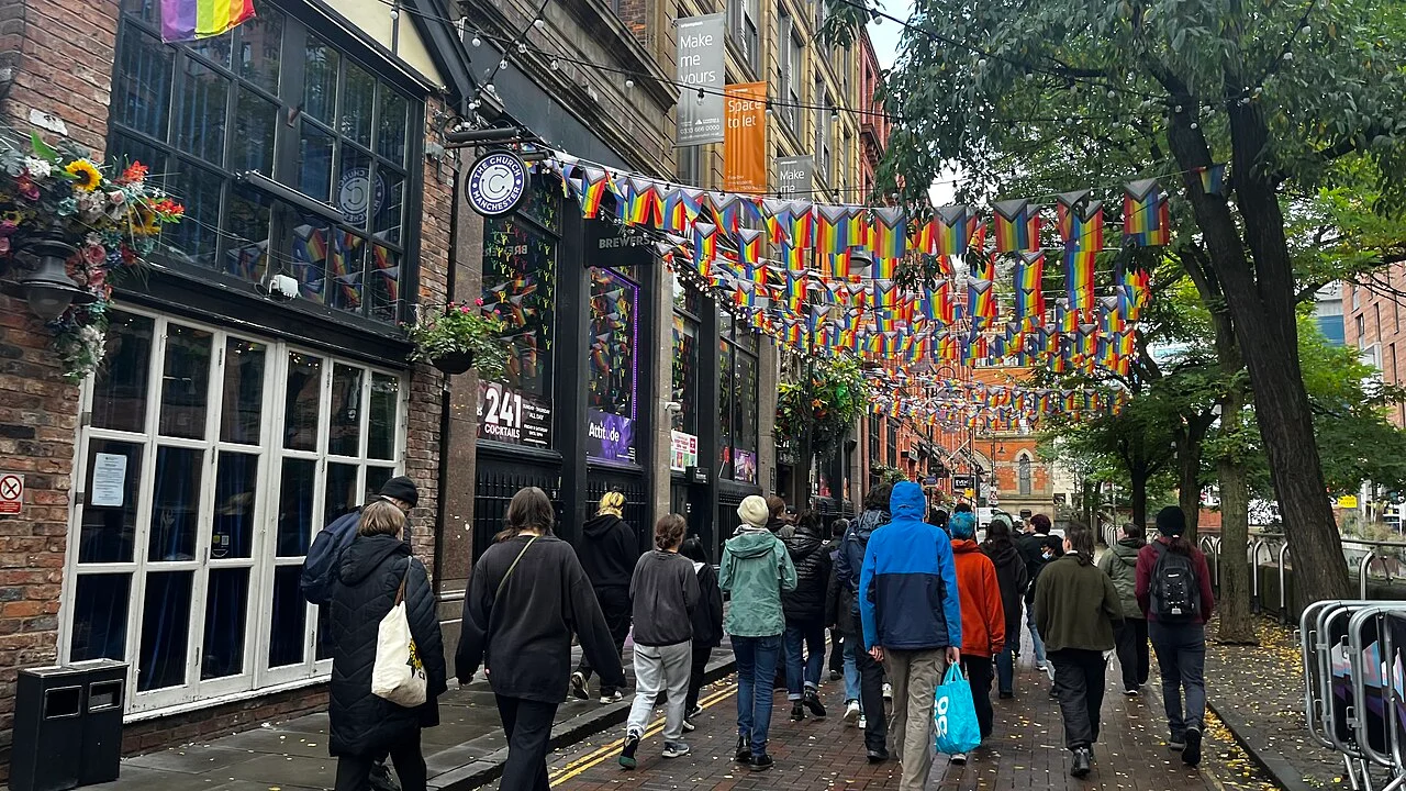 People walking along Canal Street in Manchester beneath Pride bunting, beside pub fronts and canal-side trees