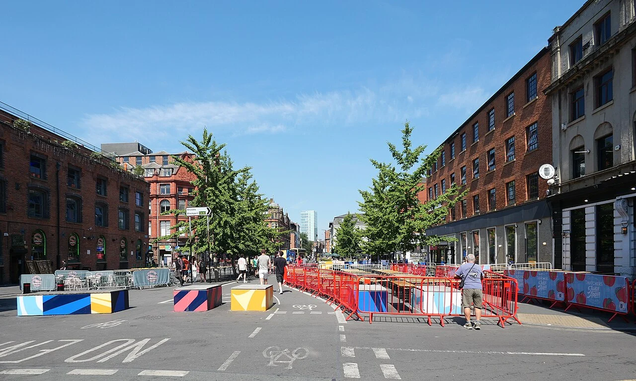 Wide view along Stevenson Square in Manchester with brick commercial buildings, trees and pedestrian street barriers