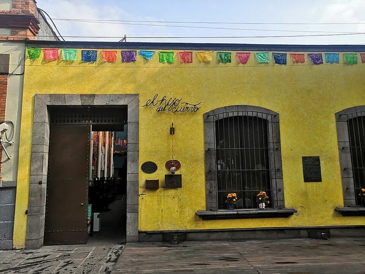 Yellow facade of El Hijo del Cuervo in Coyoacán with its script name sign, open doorway and papel picado bunting, 2019