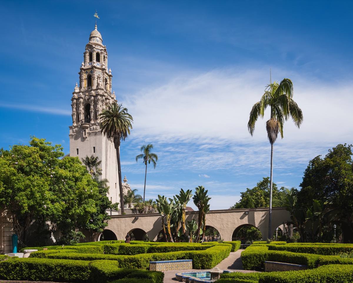 An ornate Spanish-style tower rising above clipped hedges and palm trees under a blue sky