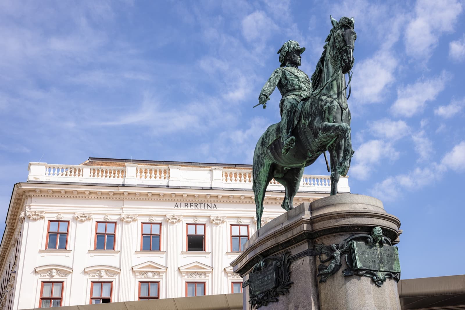 A bronze equestrian statue on a high plinth before the white palace front of the Albertina