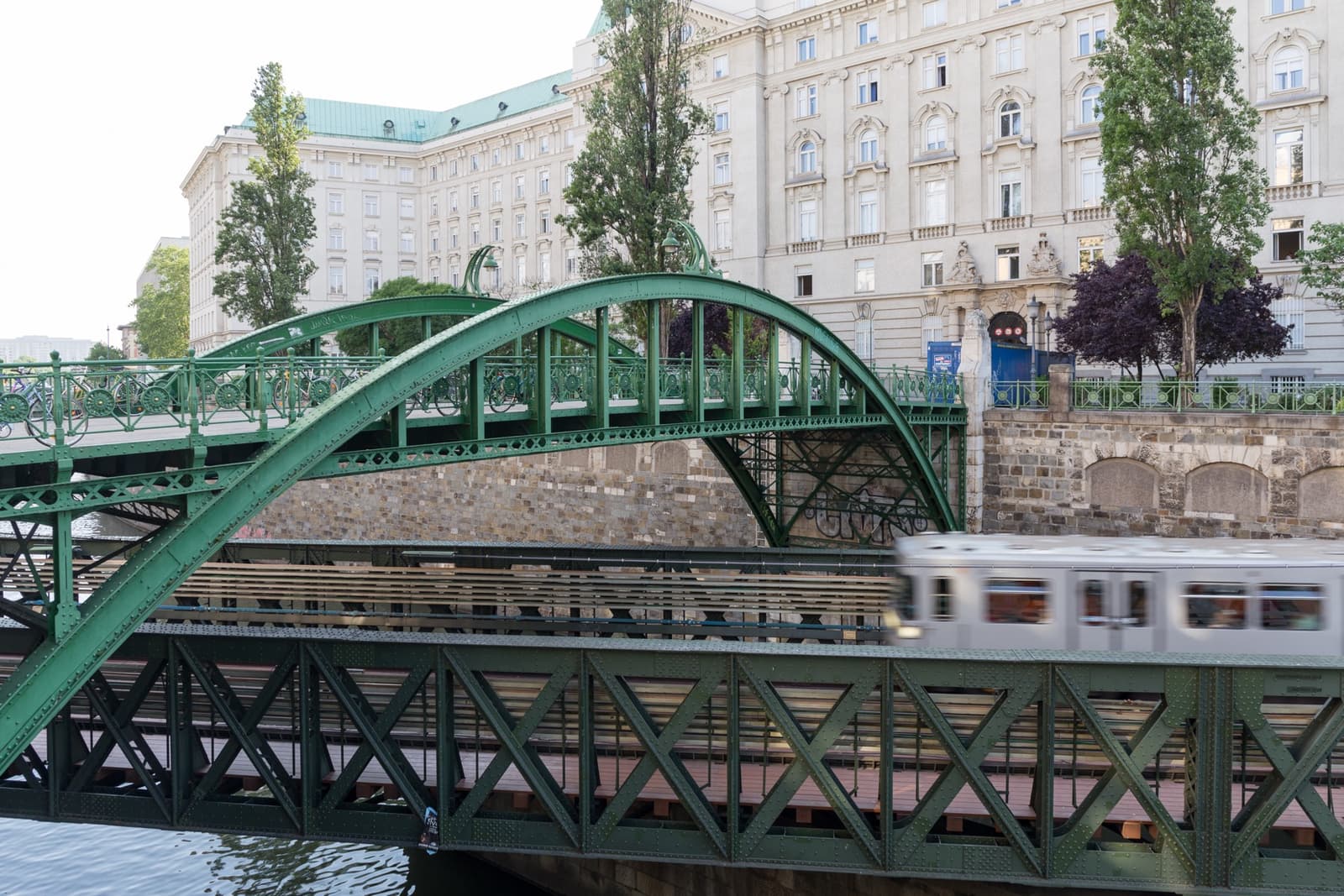 A green iron footbridge arching over a lower railway bridge where a silver train passes