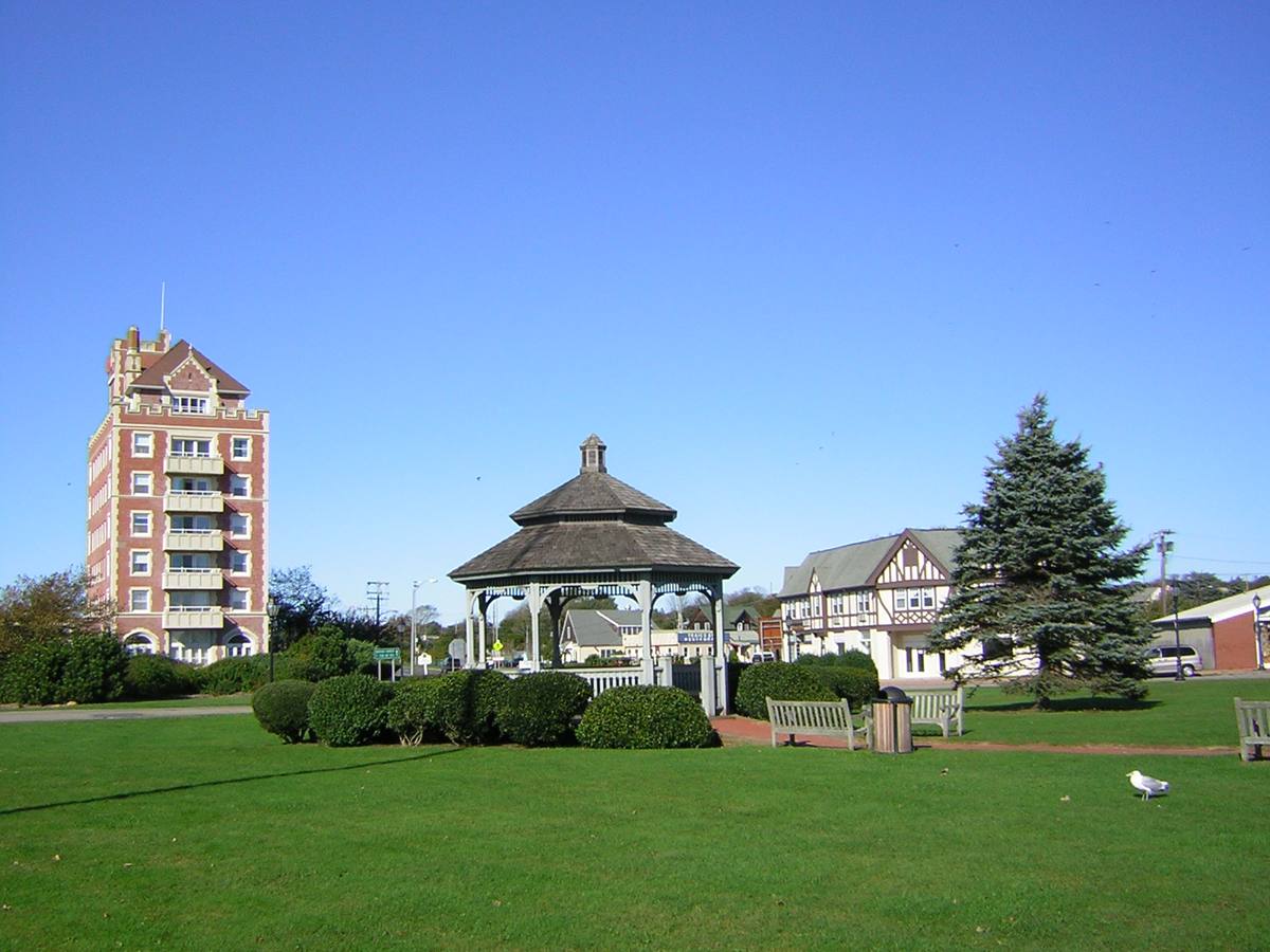 A white gazebo on a green lawn with a tall brick tower behind it