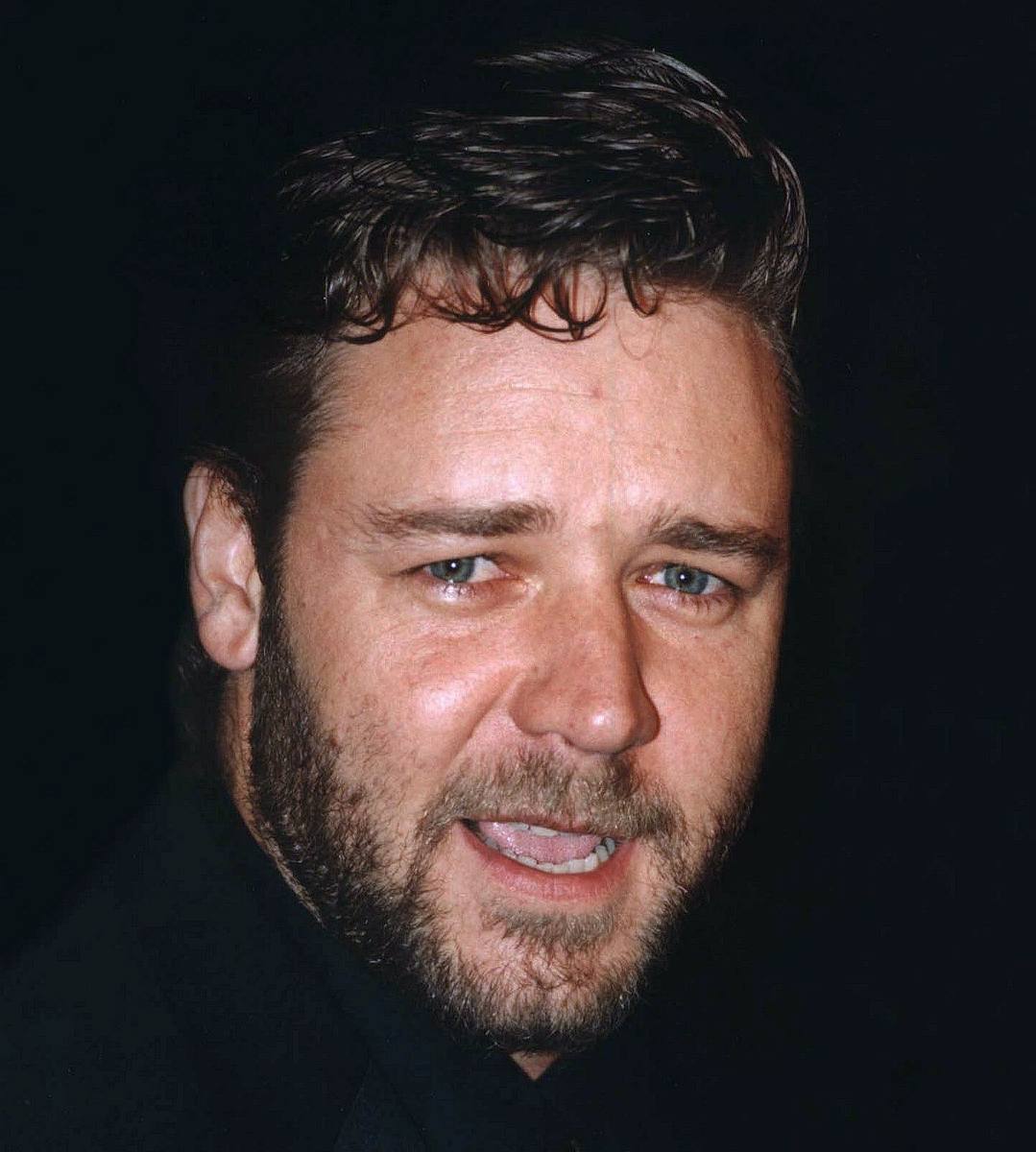 Russell Crowe, bearded, in a dark jacket, mid-word against a black background