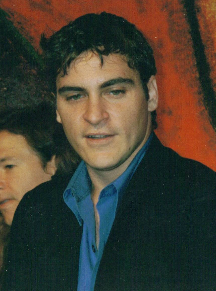 Joaquin Phoenix in a dark jacket and blue shirt in front of a red painted backdrop