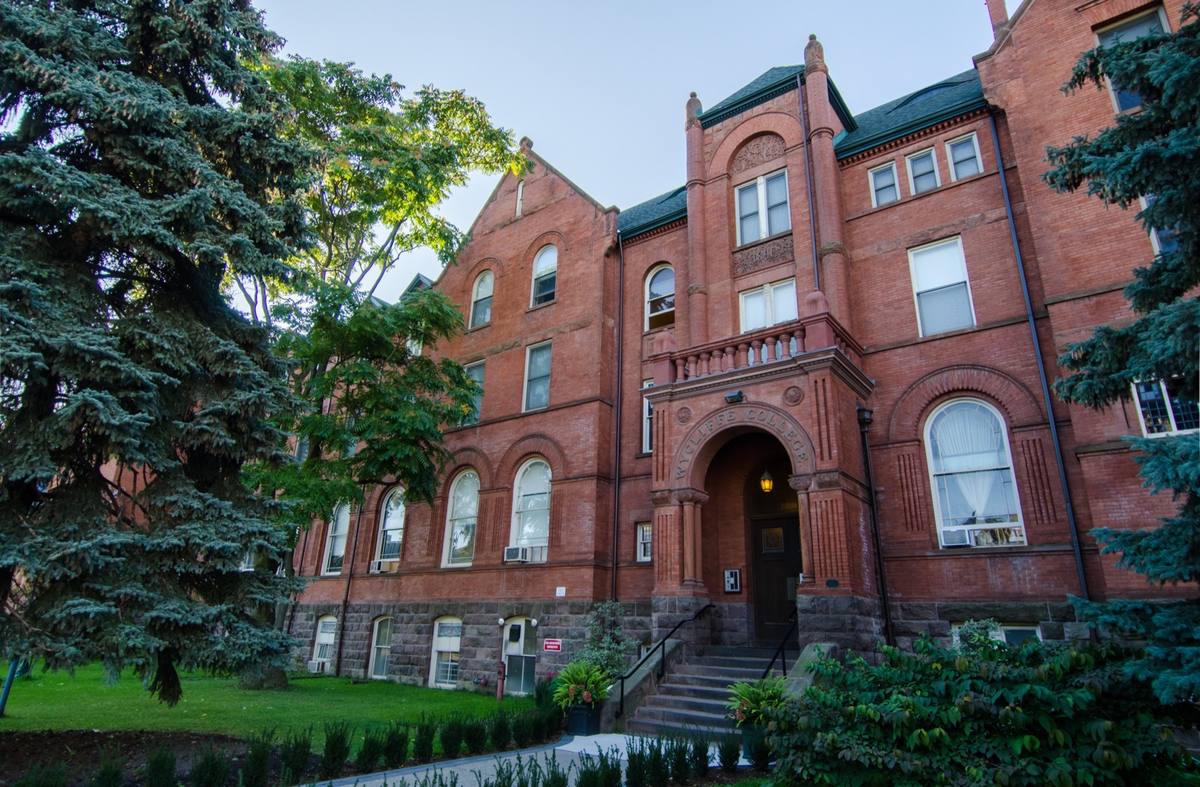 A red-brick Victorian college building with an arched entrance among fir trees