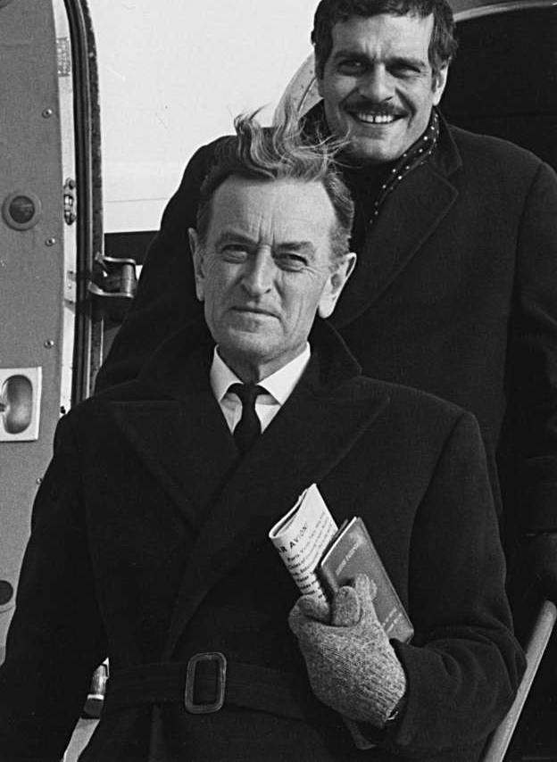 David Lean in a dark overcoat on aircraft steps, Omar Sharif smiling behind him
