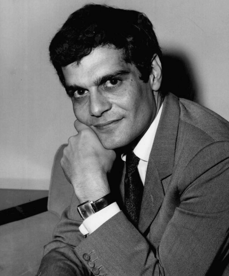 Omar Sharif in a suit and tie, chin on his hand, smiling