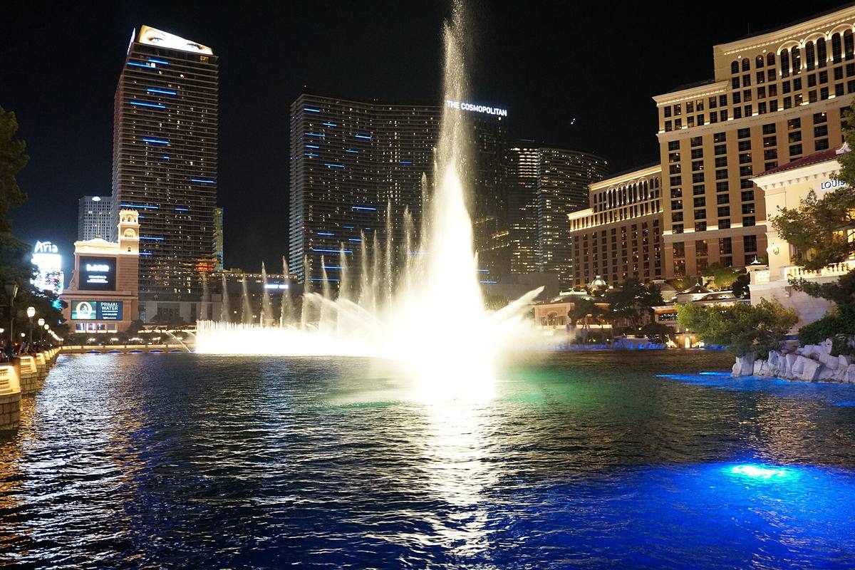 A tall white plume of water lit at night on a dark lake, hotel towers behind