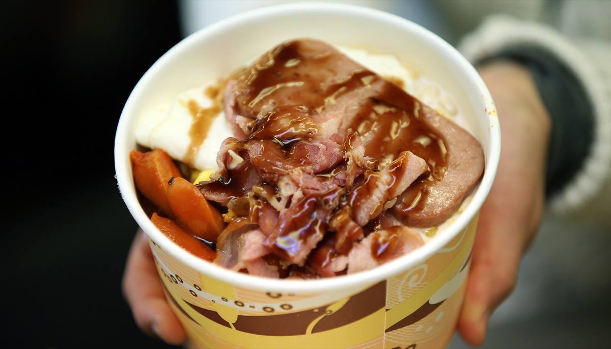 Colour photograph of a hand holding a paper cup of rice topped with meat and sauce
